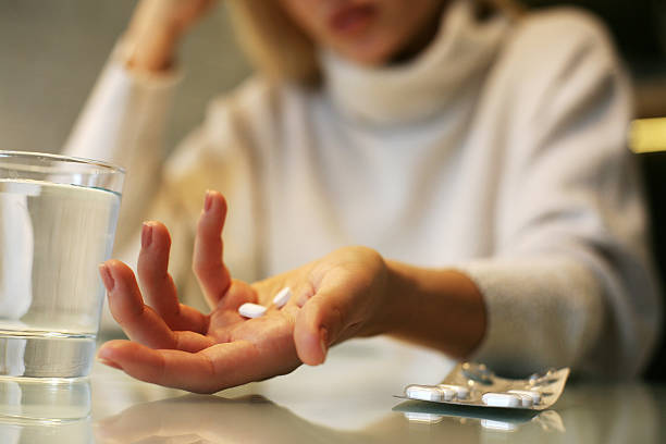 Mulher segura dois comprimidos em uma mão, em cima da mesa tem um copo de água e a cartela do medicamento | Tratamento com medicamentos para a enxaqueca