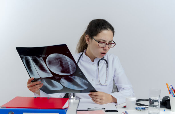 impressed young female doctor wearing medical robe and stethoscope and glasses sitting at desk with medical tools holding x-ray shot looking down isolated on white background