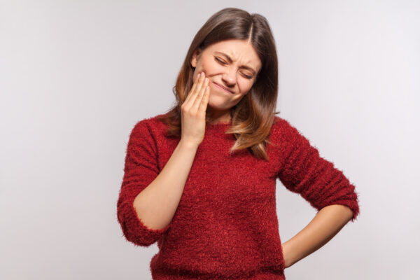 Dental problems, toothache. Portrait of unhappy girl in shaggy sweater touching sore cheek, suffering from cavities, cracked teeth, gum recession. indoor studio shot isolated on gray background