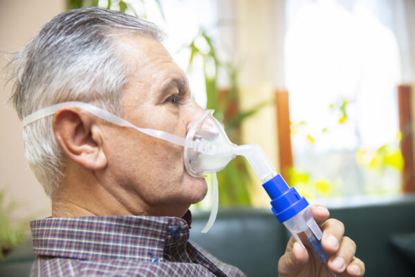 Senior man using medical equipment for inhalation with respiratory mask, nebulizer in the room
